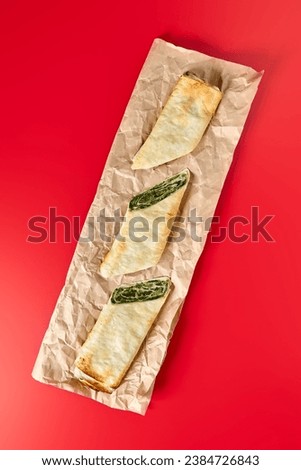 Three slices of spinach and feta cheese burek, a traditional Balkan pastry, arranged neatly on crinkled parchment paper with a bold red background.  Royalty-Free Stock Photo #2384726843
