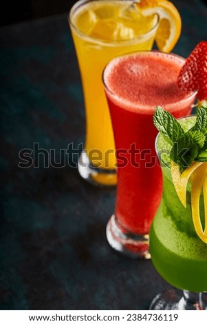 A picture of several cups of various natural juices on a green background, pineapple,  orange and strawberry juice