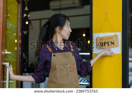 Owner of  coffee shop, small business, grabbed the door and turned and held up a welcome sign. Open shop in the morning And preparing to welcome customers, a beautiful Asian barista wears an apron.