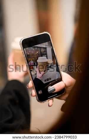 Selective focus of mobile screen with photo of paper cup takeaway coffee decorated with rose flower in female hand. Female hand taking picture of paper coffee cup