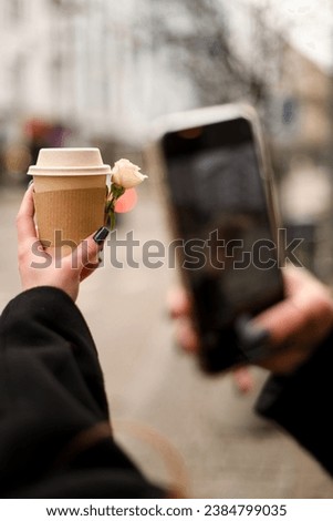Woman in warm outerwear taking photo of paper cup with takeaway drink decorated with rose flower on smartphone while standing on street. Coffee to go. Blurred background.