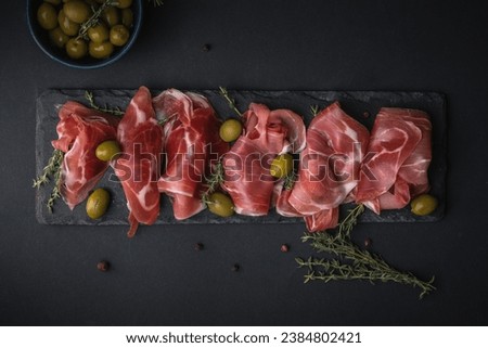 Appetizer from dry cured serrano ham or Spanish jamon iberico. Italian prosciutto crudo served with green olives on the black slate stone board. Meat slices on the black background Royalty-Free Stock Photo #2384802421