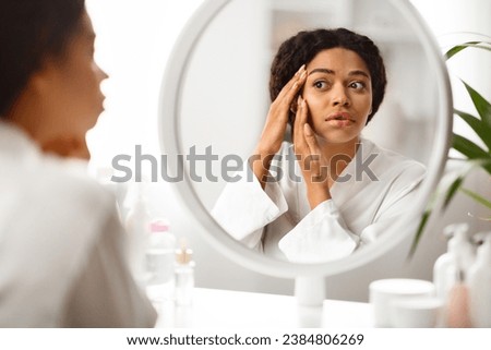 Sad african american woman looking in mirror at wrinkles around her eyes, young black lady inspecting face, feeling stressed after noticing aging signs, selective focus on reflection