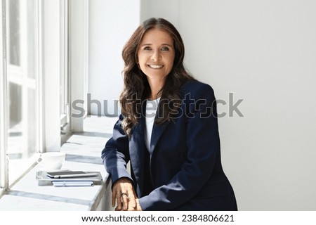 Cheerful attractive middle aged woman in formal outfit have coffee break at office, standing by window sill, smiling at camera, waiting for call from business partner, copy space Royalty-Free Stock Photo #2384806621