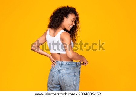 African American woman demonstrates her perfect body size after great weight loss, wearing big jeans, standing back to camera on yellow studio backdrop. Slimming motivation concept