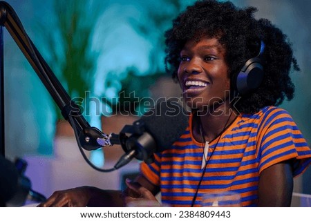 A cheerful multicultural podcaster is sitting at a recording studio with headphones on and speaking at the microphone during a podcast episode. A journalist is going live on air and live streaming