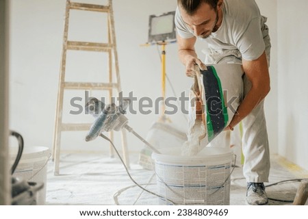 A constructor is standing in a house in a renovation process, adding plaster into a bucket and making skim coating paste. A builder pouring plaster in a bucket and preparing material for plastering. Royalty-Free Stock Photo #2384809469