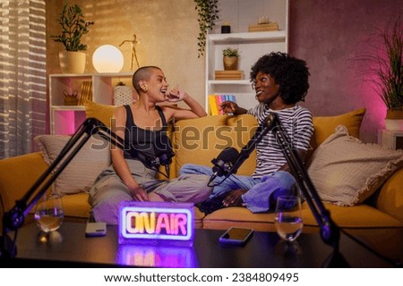 Portrait of diverse women recording a podcast while using a microphone and sitting on a sofa in a modern room. Female podcaster making audio podcast from her home studio and interviewing her guest.