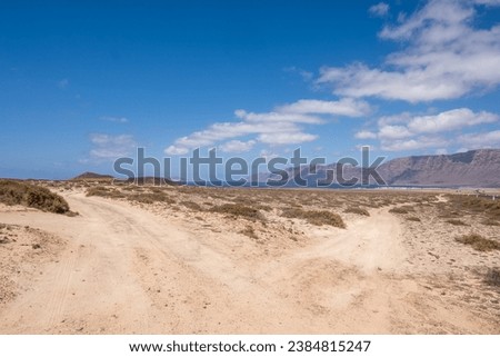 Desert landscape of white sand and desert bushes. Ocean and Famara cliff in the background. Dirt track. Sky with big white clouds. Lanzarote, Canary Islands, Spain.