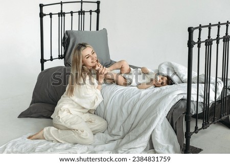 A little girl lies on the bed and drinks water from a bottle, next to her mother kisses her feet. Family concept. View through the metal bars of the bed.
