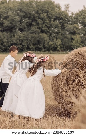 Children push a bale of hay. Twin girls in white dresses with their brother. Photoshoot. Family.