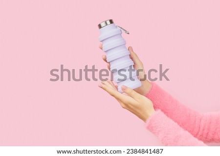 Womans hands holding collapsible reusable lilac water bottle on pink background. Sustainable lifestyle