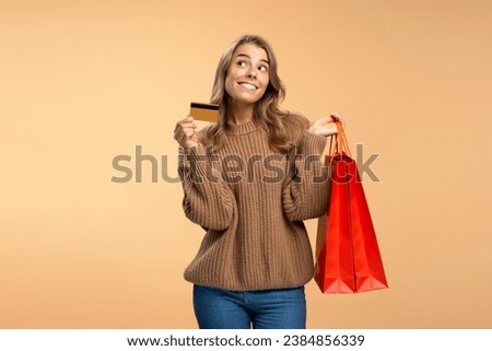 Attractive smiling woman holding credit card and red shopping bags looking away choosing clothing isolated on beige background, copy space. Shopping, sale, black Friday, advertisement concept
