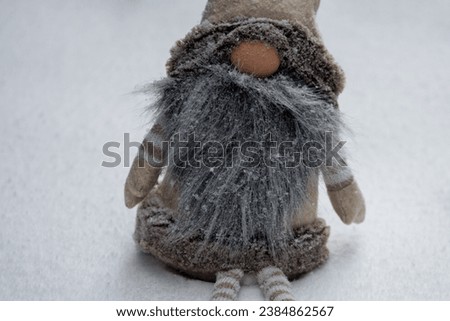 A small stuffed gnome toy on white snow. The little traditional scandinavian mythical doll has a thick grey beard, large beige nose and a large hat. Its coat and legs are stripped brown and beige wool