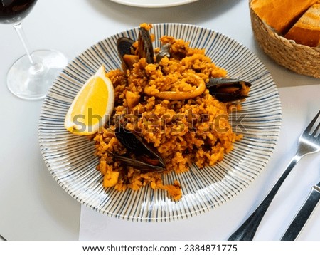 Appetizing racy seafood paella with mussels and squid rings served with lemon slice and glass of red wine. Traditional Spanish cuisine. Royalty-Free Stock Photo #2384871775