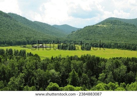 A huge clearing with a large farm surrounded by a fence at the foot of a high mountain with a dense coniferous forest on a cloudy summer day. Khakassia, Siberia, Russia.