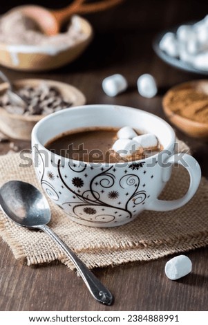 A tea cup filled with hot cocoa and marshmallows with ingredients in behind.
