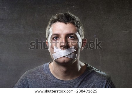 attractive young man with mouth sealed on duct tape to prevent him from speaking keeping him mute and censored in freedom of speech and expression concept isolated on dark grunge studio light style Royalty-Free Stock Photo #238491625