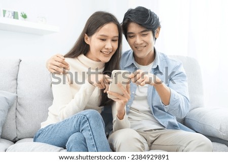Happy couple love at home, two asian young spending time, bonding to each other romantic on sofa in living room while man embrace woman using smartphone, mobile phone to selfie take a photo together.