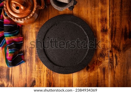Empty stone comal plate on wooden rustic table, colorful typical mexican fabric with clay pot and stone molcajete, traditional kitchen utensil used in Mexican gastronomy. Royalty-Free Stock Photo #2384934369