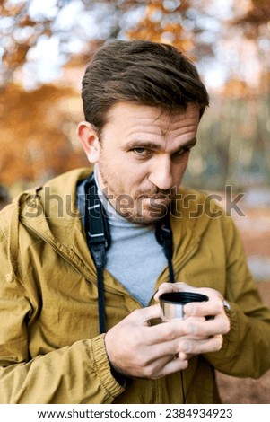 Young man with a cup of coffee in his hands looks frowningly while standing in the park