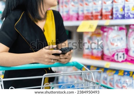 Woman using smartphone with trolley cart while walking and taking products from shelf in a grocery store.