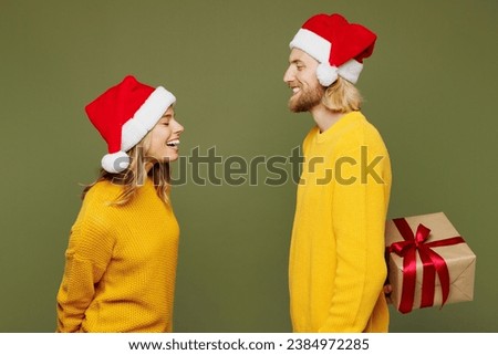 Side view merry young couple two friends man woman wears sweater Santa hat posing give present box with gift ribbon bow isolated on plain green background. Happy New Year celebration Christmas concept