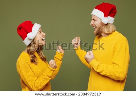 Sideways merry young couple two friends man woman wear sweater Santa hat posing do winner gesture clench fist isolated on plain green background. Happy New Year celebration Christmas holiday concept