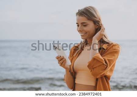 Side view young smiling calm fun woman she wearing orange shirt casual clothes hold in hand use mobile cell phone walk on sea ocean sand shore beach outdoor seaside in summer day. Lifestyle concept Royalty-Free Stock Photo #2384972401