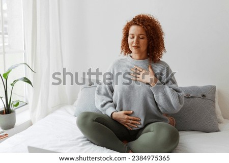 Portrait of plus-size curly redhead female in casual clothes doing pranayama technique sitting on bed next to window with closed eyes, putting hands on chest, breathing slowly and deeply Royalty-Free Stock Photo #2384975365