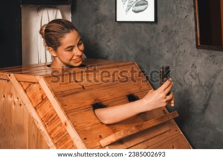 A portrait of gorgeous caucasian woman playing her mobile phone while using wooden sauna cabinet in warm tone. Attractive female with beautiful skin taking a photo. Gray background. Tranquility