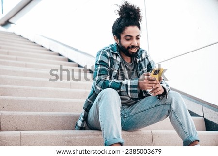 Handsome smiling hipster model. Unshaven Arabian man dressed in summer casual clothes. Fashion male with long curly hairstyle posing in the street. Holds smartphone, uses phone mobile apps