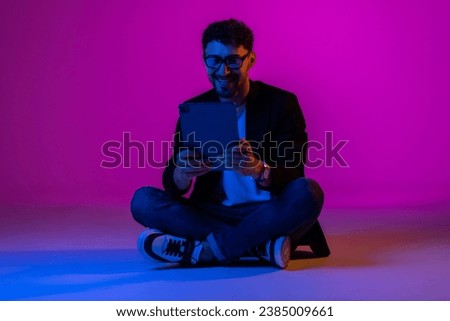 Young man, manager wearing business suit and eyeglasses using digital tablet on gradient blue purple background in neon light. Studying, online work, job, finance, new app concept