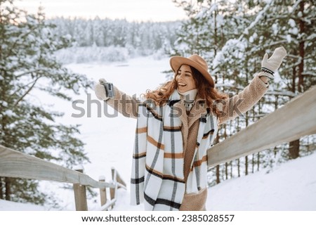 Happy woman in a hat and scarf with a phone in her hands taking a selfie in a snowy forest. Young tourist woman with phone enjoying snowy winter day near lake. Active lifestyle.