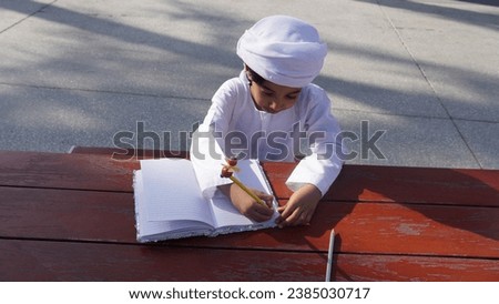Happy Arab Middle Eastern boy wearing Ghutra sitting at table learning, writing in note book.