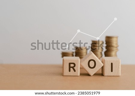 Cube block wood letters ROI and blurred money coin stack with white increasing graph including copy space. For Return on Investment financial concept Royalty-Free Stock Photo #2385051979