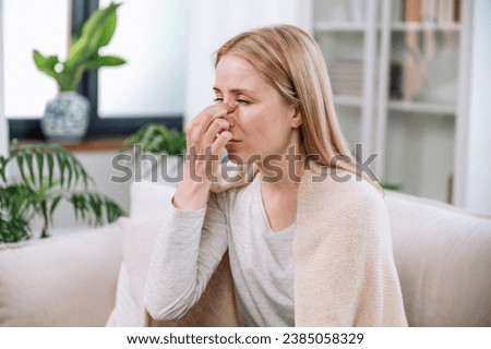 tired young woman feeling nasal discomfort touching nose and suffering from seasonal allergy while sitting on couch at home. sick female with sneezing and grippe symptoms covered in plaid on sofa Royalty-Free Stock Photo #2385058329