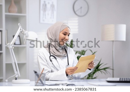 Joyful female doctor in hijab received envelope message mail notification with good results of patient's treatment, woman is happy and smiling, working inside clinic office in white medical coat. Royalty-Free Stock Photo #2385067831