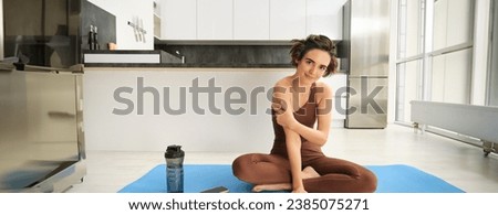Sport and wellbeing. Young brunette fitness girl, wearing activewear, sitting on yoga mat, meditating, practice relaxation and mindfulness at home, workout in kitchen. Copy space Royalty-Free Stock Photo #2385075271