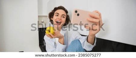 Beautiful woman eating an apple and recording herself on video. Cute girl with fruit, posing for photo, taking selfie in kitchen in happy mood.