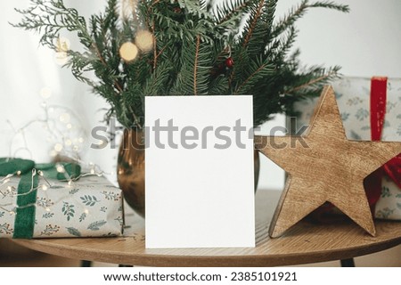 Empty greeting card, stylish wrapped christmas gift, rustic star and fir branches with lights on wooden table. Christmas card mock up. Space for text. Season greetings postcard template