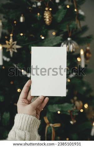 Christmas card mock up. Hand holding empty greeting card on background of stylish decorated christmas tree with golden lights. Space for text. Season greetings template and vintage ornaments Royalty-Free Stock Photo #2385101931