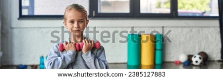 preadolescent cute blonde girl exercising with dumbbells and looking at camera, child sport, banner