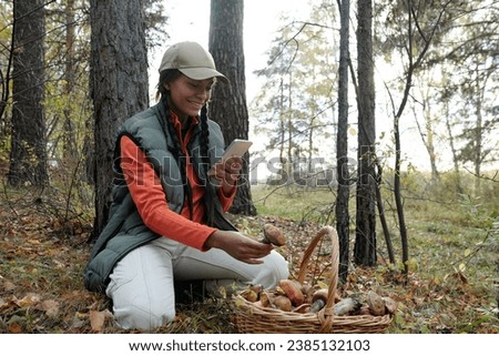 Happy young African American female blogger taking photo of picked mushrooms in basket or making video for her subscribers