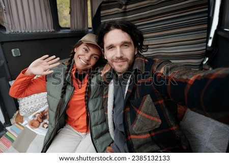 Happy young intercultural couple greeting subscribers of their channel while making livestream from travel van after picking mushrooms