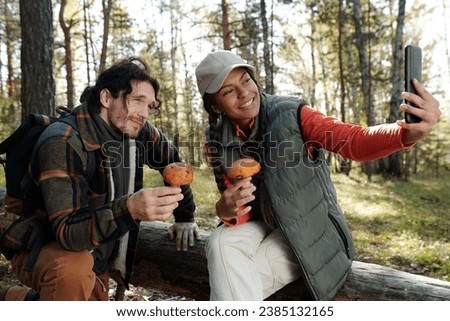 Happy young woman with smartphone and orange cap boletus mushroom taking selfie with her husband holding porcini in autumn forest