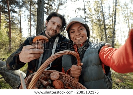 Couple of young smiling mushroom pickers with basket full of various fungi looking at camera while taking selfie in autumn forest