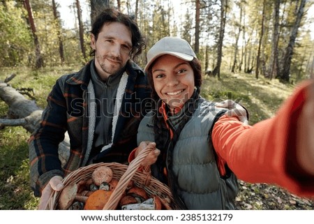 Young intercultural couple of mushroom pickers taking picture on mobile phone while both looking at camera in autumn forest