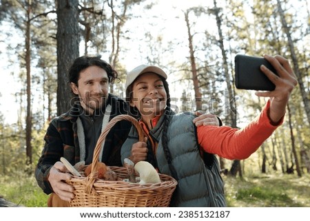 Young intercultural dates with basket of mushrooms looking at smartphone camera with smiles during selfie in autumn forest