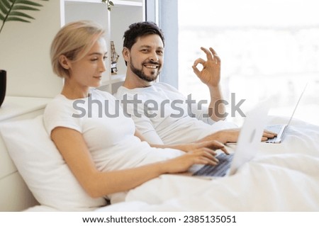 Caucasian male scrolling through webpages on laptop in bed showing sign ok while busy lady typing on portable computer on background. People in relationships spending quality time together at home.